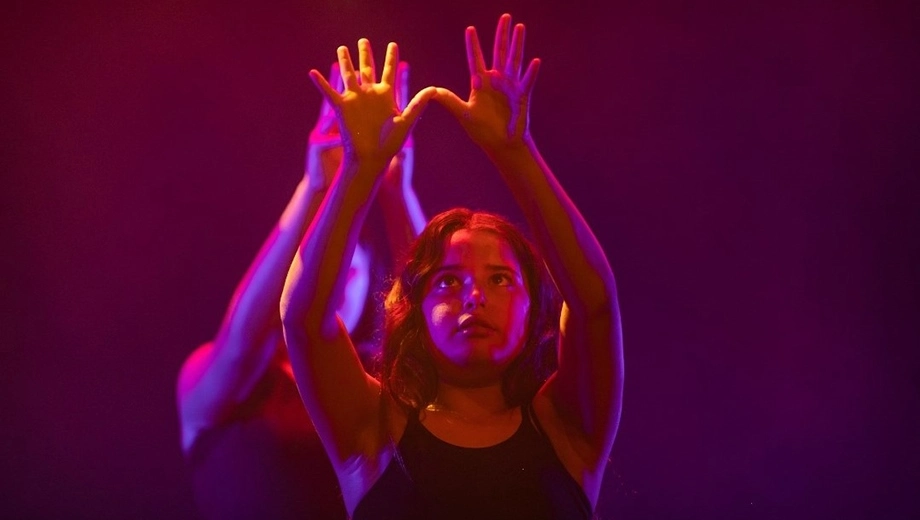 Two performers on stage with their arms raised, illuminated by coloured stage lighting.